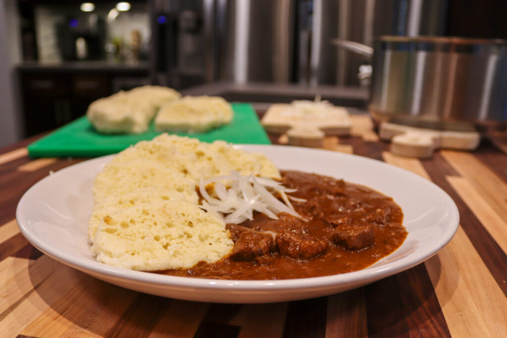 Czech Beef Goulash with Dumplings and fresh onion side shot with extra dumplings in the background, kitchen in the background