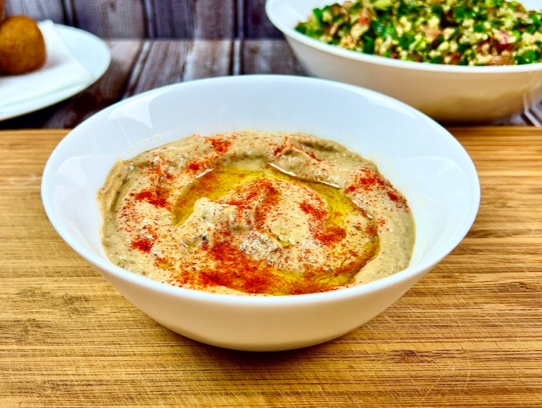 Creamy & Smoky Baba Ganoush in the white bowl on the light brown countertop, vegetable in the background.