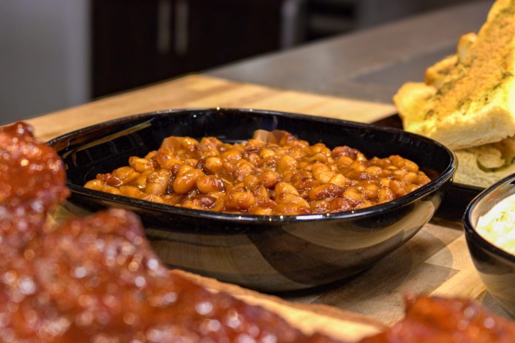 Easy Baked Beans in black bowl on the butcher block countertop in medium distance shot serrounded by pork ribs, coleslaw, and baked baguette