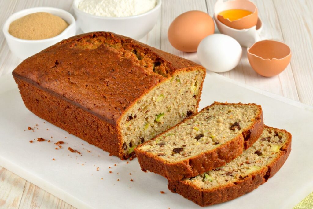 Classic Zucchini Bread recipe on the cutting board, partially cut, serrounded by som of the ingredients, side medium distance shot