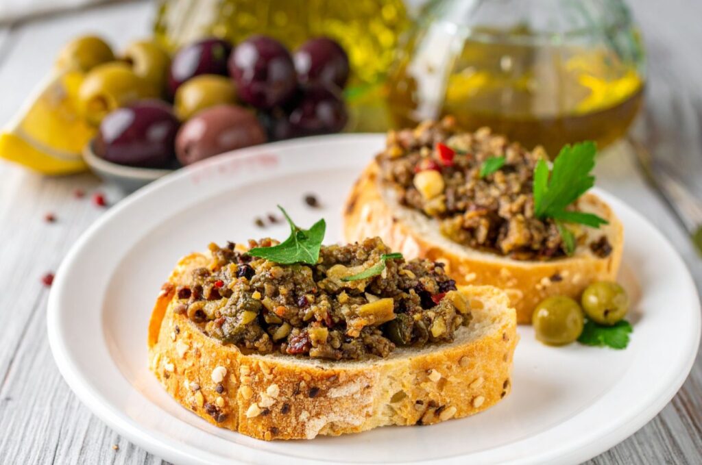 Olive Tapenade Recipe on the fresh slices of bread with other ingredients in the background
