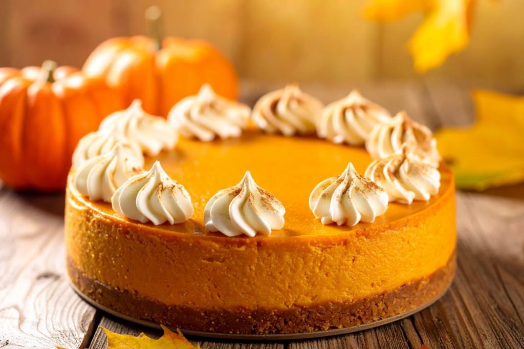 Pumpkin cheesecake decorated with whipped cream on the cutting board on the rustic table with pumpkins in the background. Side medium distance shot.